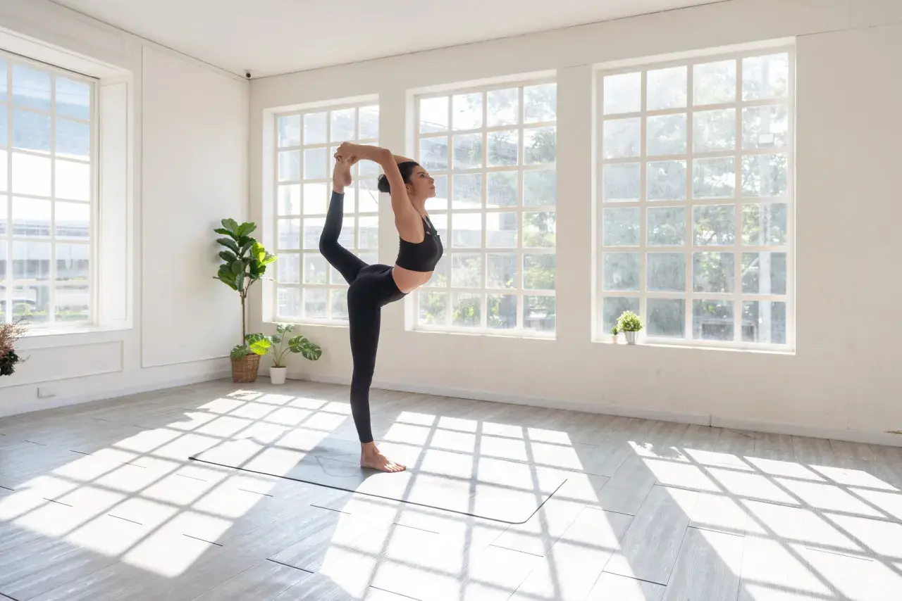 Kobieta w czarnym stroju wykonuje pozycję Natarajasana podczas sesji vinyasa jogi. Światło wpada przez okna.