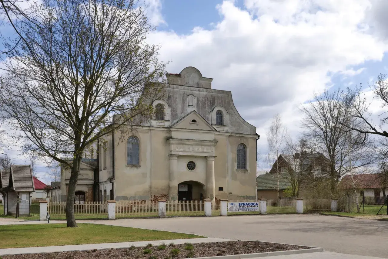 Synagoga w Orli zaprasza. Stary budynek z charakterystycznym dachem i oknami, otoczony płotem i drzewami.