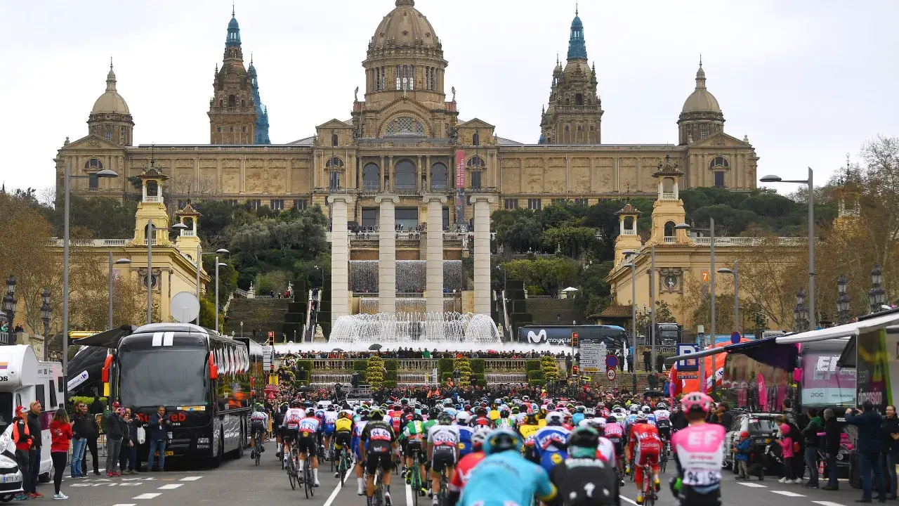 Kolarze w barwnych strojach pędzą ulicą przed monumentalnym budynkiem z fontannami, przygotowując się do etapu Tour de France.
