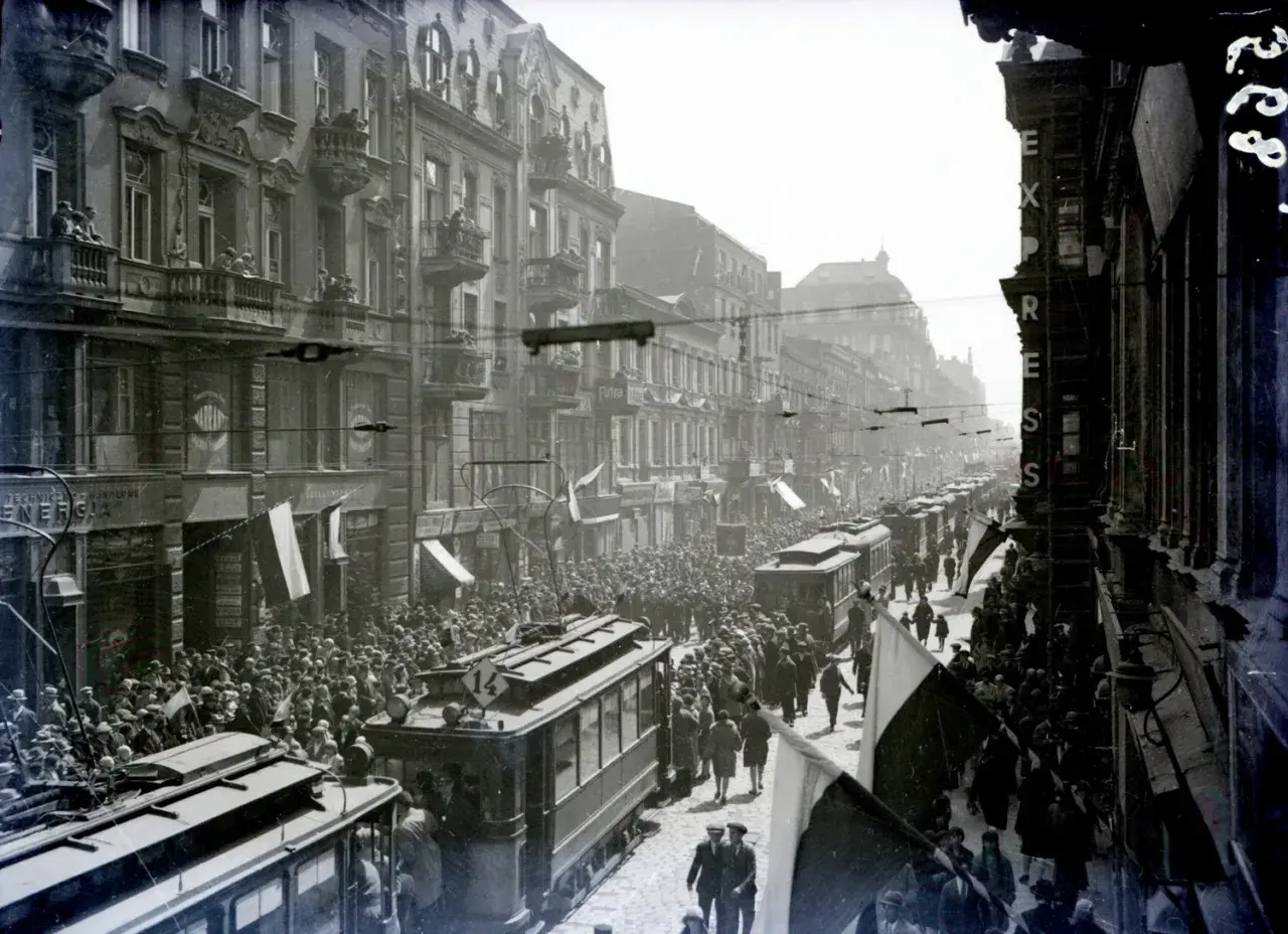 Tłum ludzi i tramwaje na ulicy Piotrkowska w Łodzi. Widać kamienice, flagi i napisy na sklepach.
