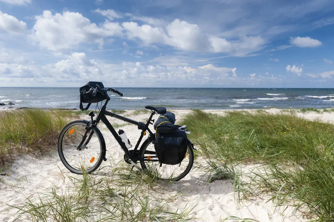 Rower z sakwami na piaszczystej plaży, gotowy na odkrywanie tras rowerowych nad morzem. Błękitne niebo i fale tworzą idealne tło.
