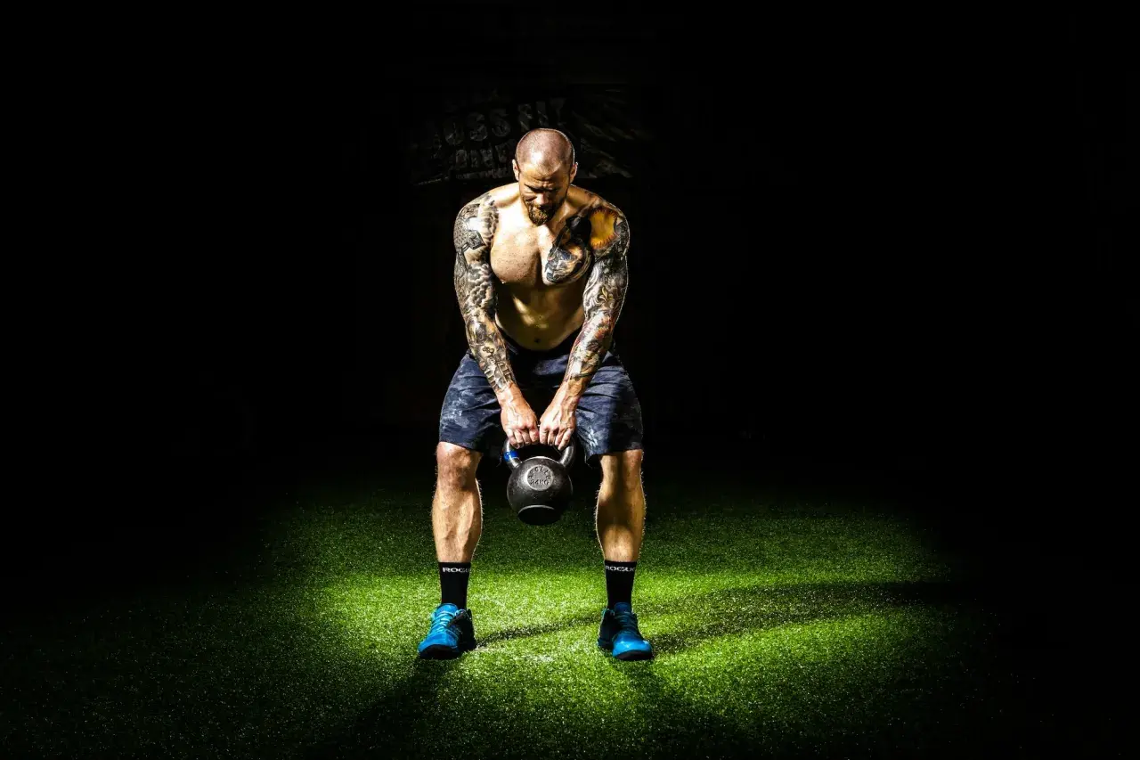 Mężczyzna z tatuażami trzyma kettlebell na tle ciemności i zielonej trawy.