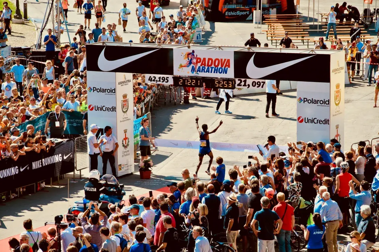 Biegacz przekracza metę maratonu w Padwie, świętując zwycięstwo. Tłum kibiców i zawodników dopingują na trasie.
