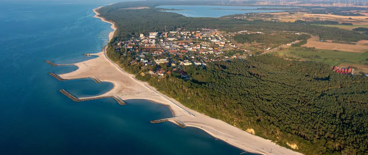 Widok z lotu ptaka na Jarosławiec, nadmorską miejscowość z piaszczystą plażą, lasami i zabudowaniami.