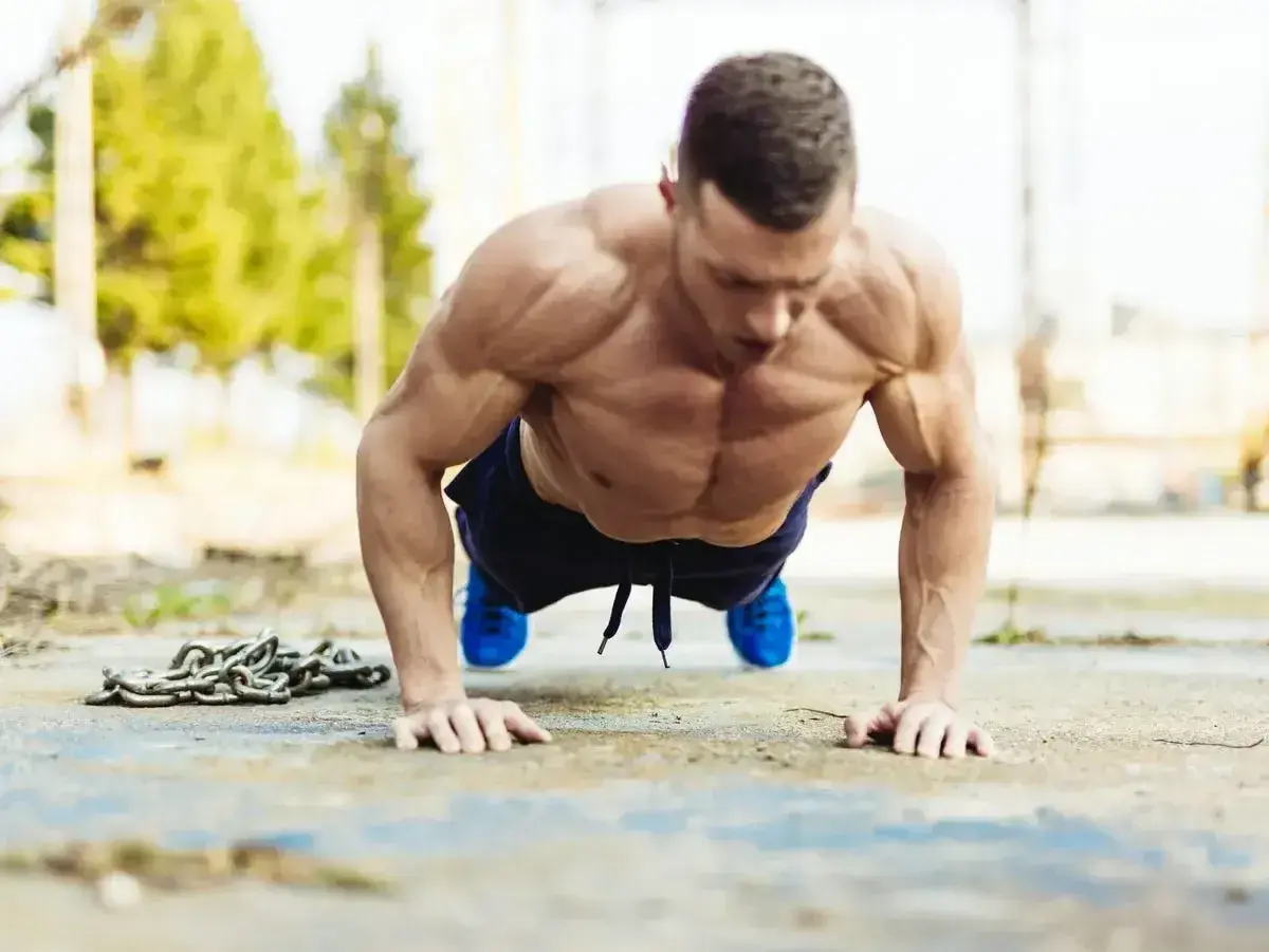 Mięśniowy mężczyzna wykonuje pompki na biceps na zewnątrz. Obok leży ciężki łańcuch, symbolizujący trudny trening.