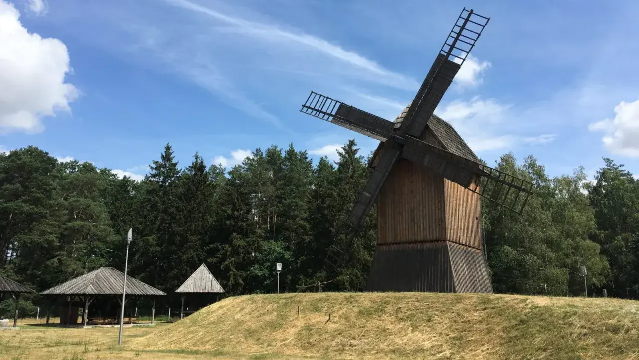 Drewniany wiatrak na wzgórzu, otoczony lasem, w tle błękitne niebo z chmurami. Miejsce jak z bajki w Obornikach Śląskich.