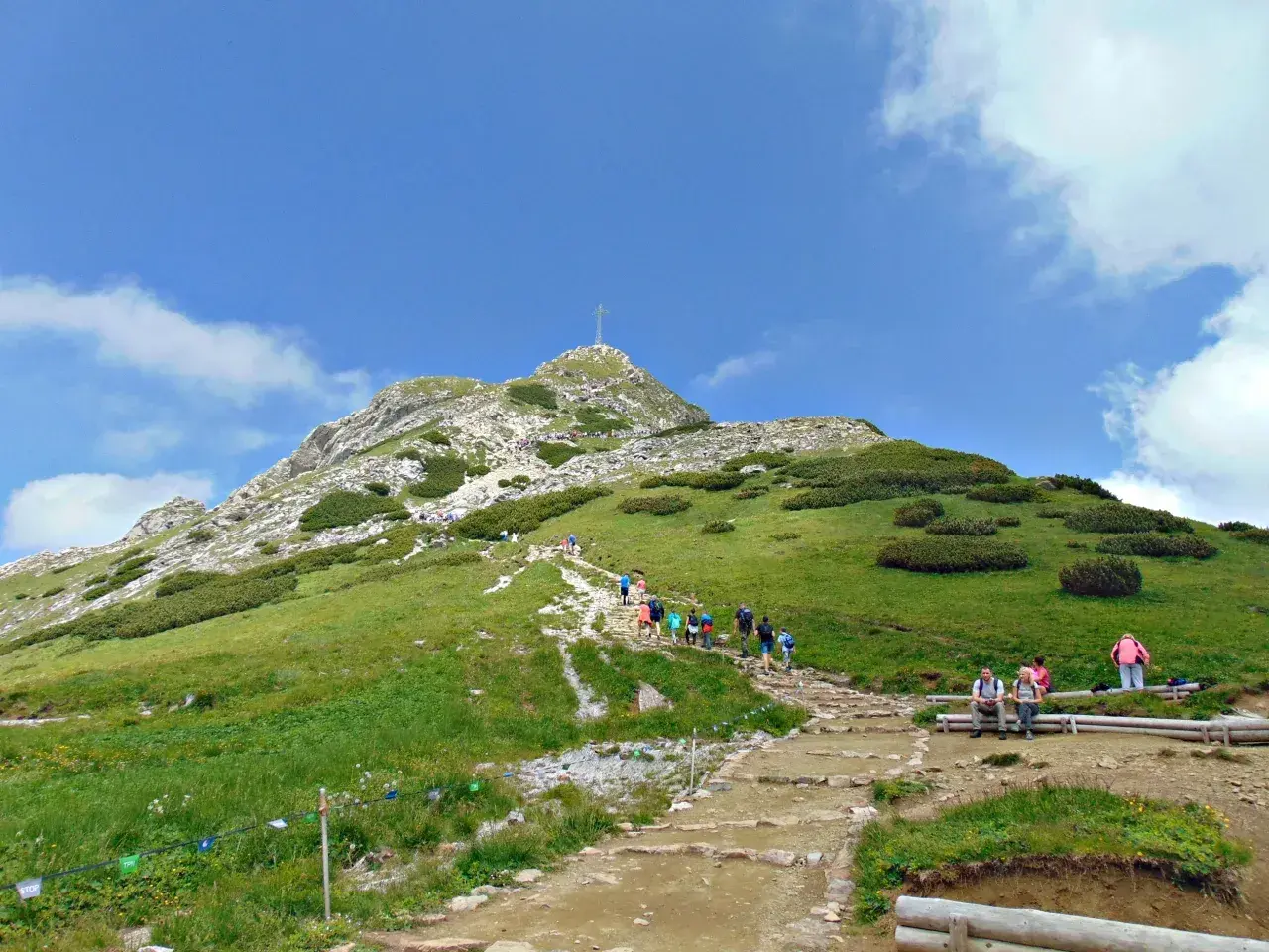 Tłum turystów wspina się kamienistym szlakiem na Giewont. Widok na szczyt z krzyżem i zielone zbocza Tatr.