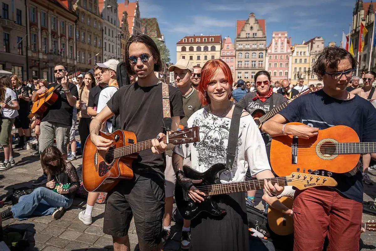 Młodzi muzycy grają na gitarach na wrocławskim Rynku. W tle kolorowe kamienice i tłum ludzi.