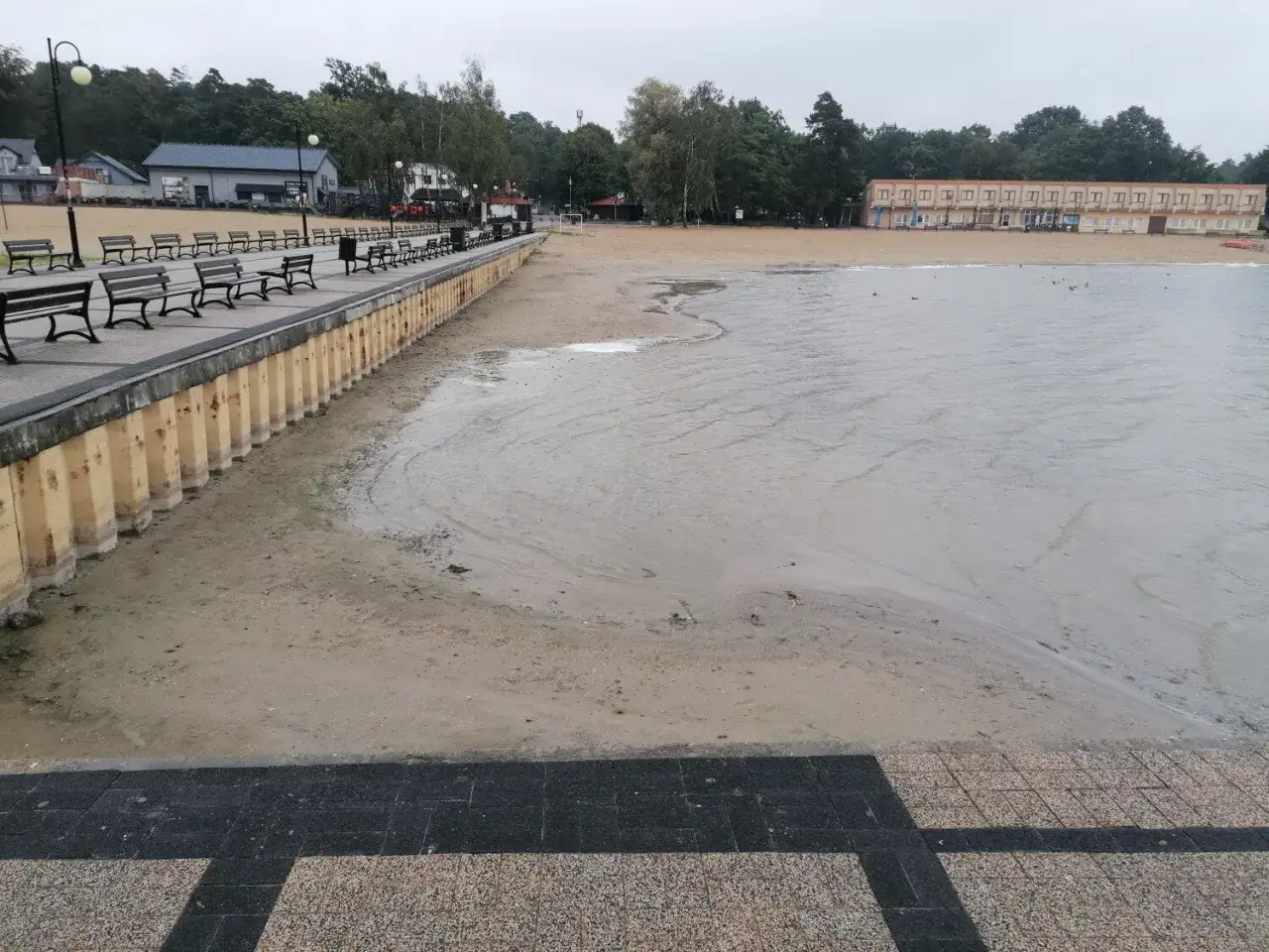 Skorzęcin atrakcje: pusta plaża z ławkami i budynkami w tle, spokojna woda.