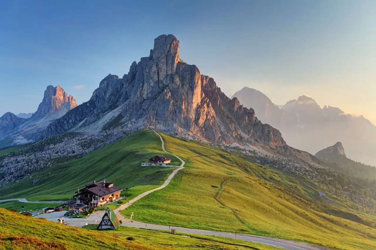 Dolomity o wschodzie słońca. Szlak Alta Via 1 wije się przez zielone wzgórza do schroniska, a majestatyczne szczyty górują w oddali.