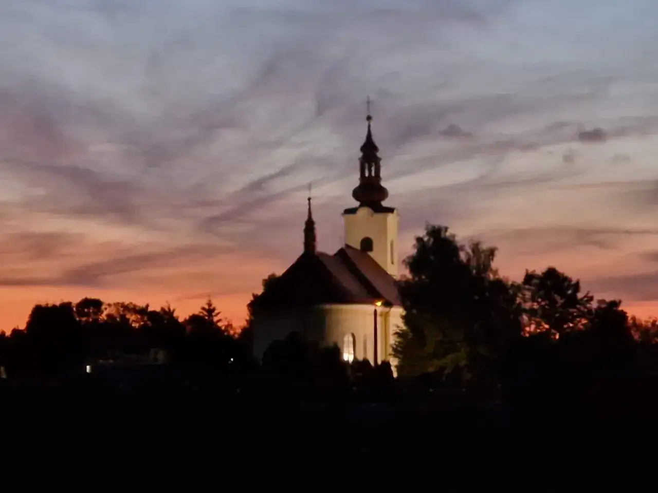 Kościół z wieżą i krzyżem na tle zachodzącego słońca. W oddali drzewa, a na niebie chmury. Włosienica w barwach pomarańczy i fioletu.