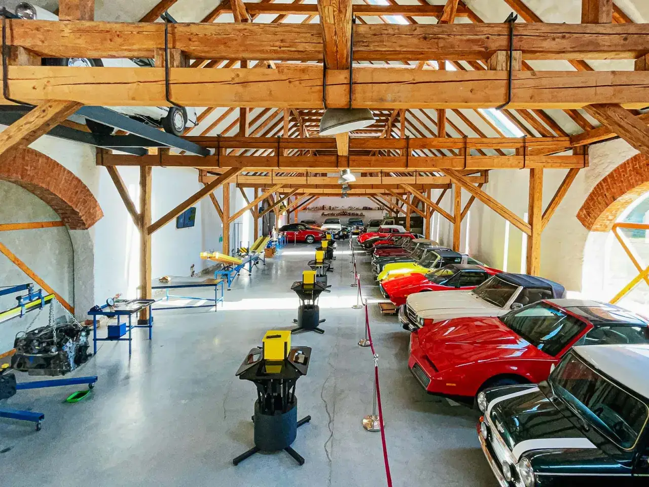 W muzeum motoryzacji, w drewnianej stodole, stoją rzędy klasycznych samochodów. Widać tu m.in. czerwone, żółte i białe modele, a także czarny Mini Cooper. W tle widać podnośnik z silnikiem, co sugeruje, że to miejsce to nie tylko ekspozycja, ale też wa...