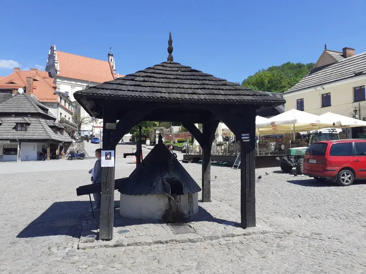 Rynek w Kazimierzu Dolnym: zabytkowa studnia pod drewnianą altaną, kamienne bruki, budynki z czerwonymi dachami i traktorem.