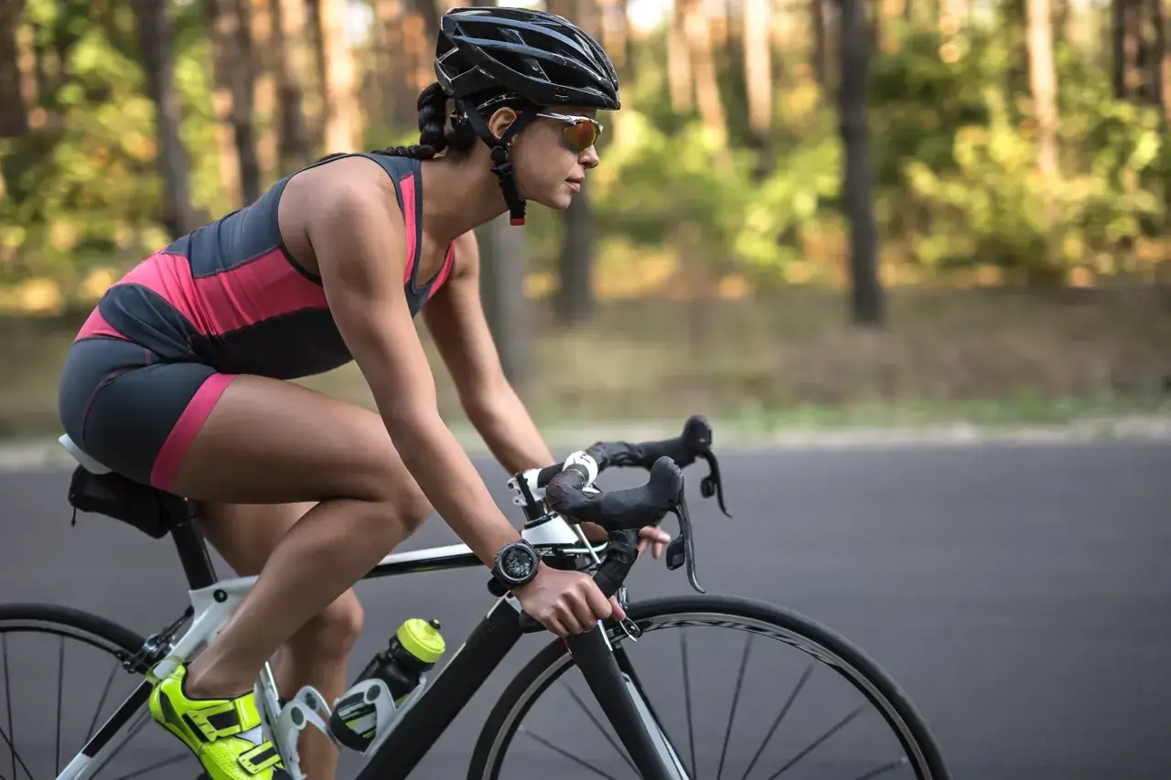 Kobieta w kasku i okularach jedzie na rowerze szosowym przez las. Widać jej nogi w sportowym stroju i zegarek na ręce. Jazda na rowerze - efekty po miesiącu są widoczne w jej sylwetce.