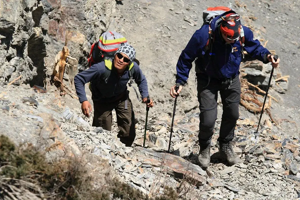 Dwóch turystów z plecakami wspina się po kamienistym zboczu, używając kijków do trekkingu. Chodzenie z kijkami ułatwia im pokonywanie trudnego terenu.