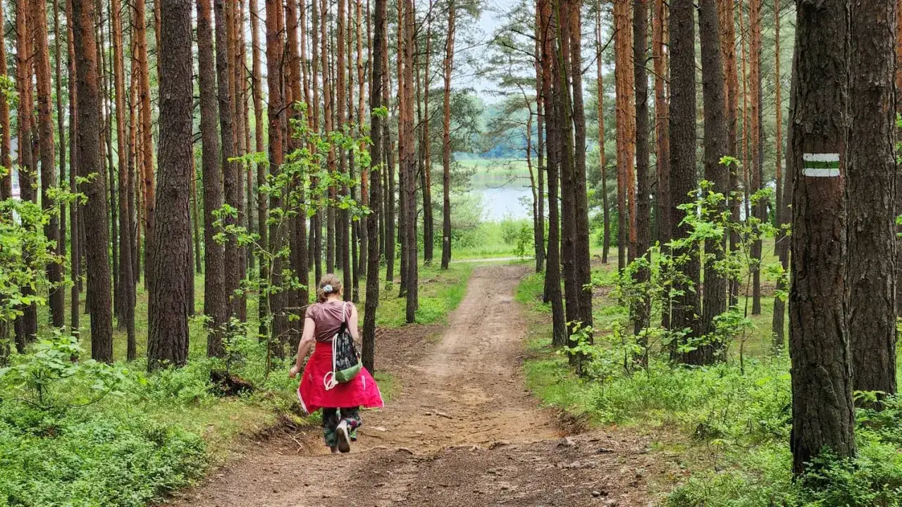 Kobieta w czerwonej spódnicy idzie leśną ścieżką w kierunku jeziora. Szlak turystyczny w lesie Poraj.