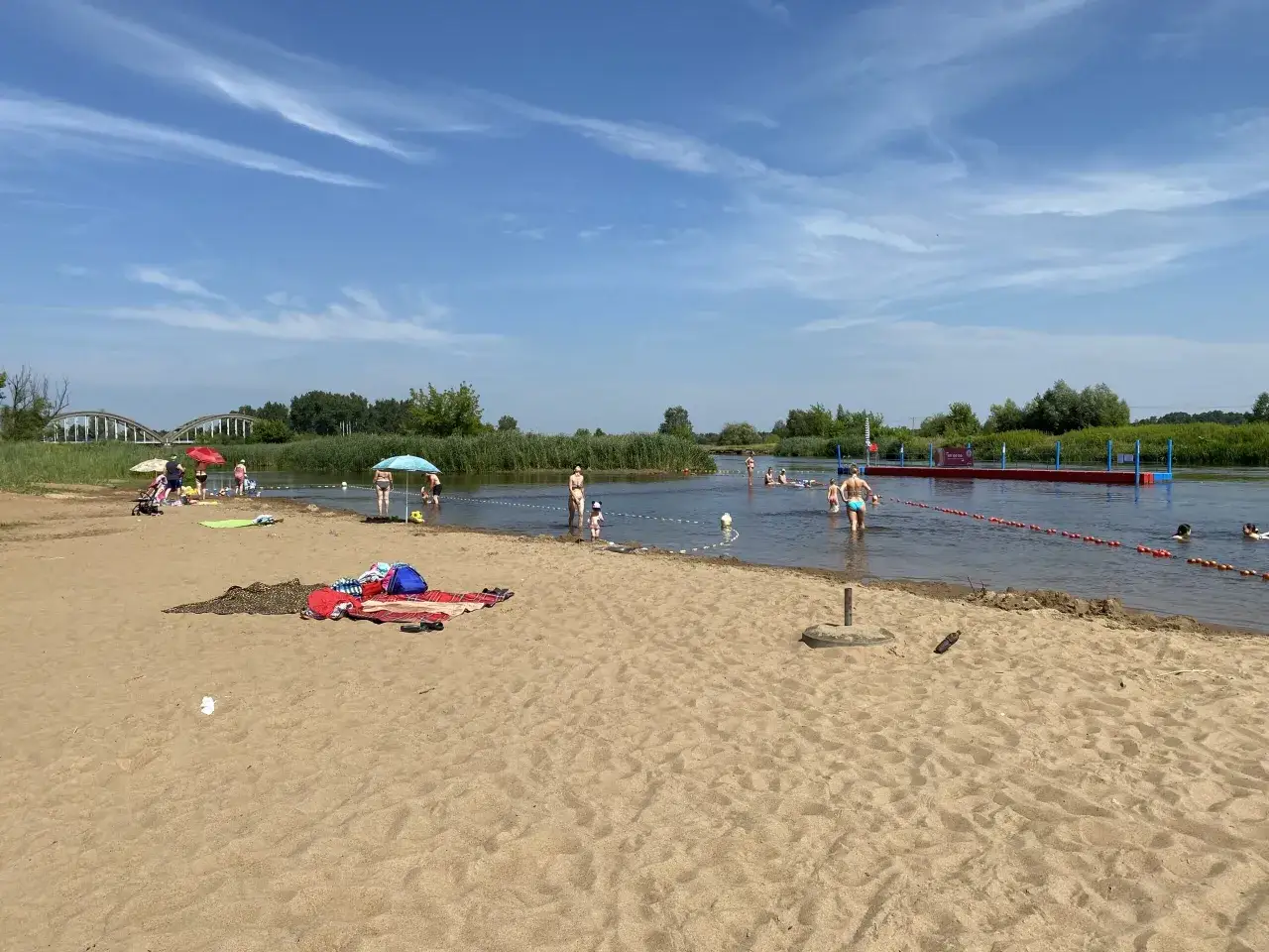 Ludzie odpoczywają na piaszczystej plaży, niektórzy kąpią się w rzece. W oddali widać most i zielone drzewa. To idealne miejsce na letni relaks nad wodą, z piaszczystym białobrzegiem.