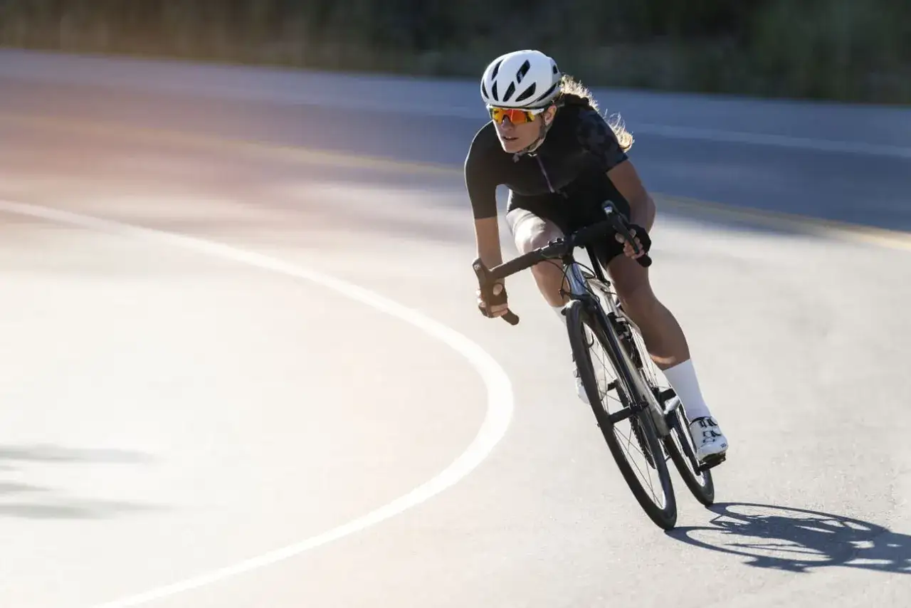 Kobieta w kasku i okularach ściga się na rowerze szosowym, pochylona w zakręcie. Widać jej sportowy strój i białe skarpetki. To prawdziwa pasja do bikeryzmu.