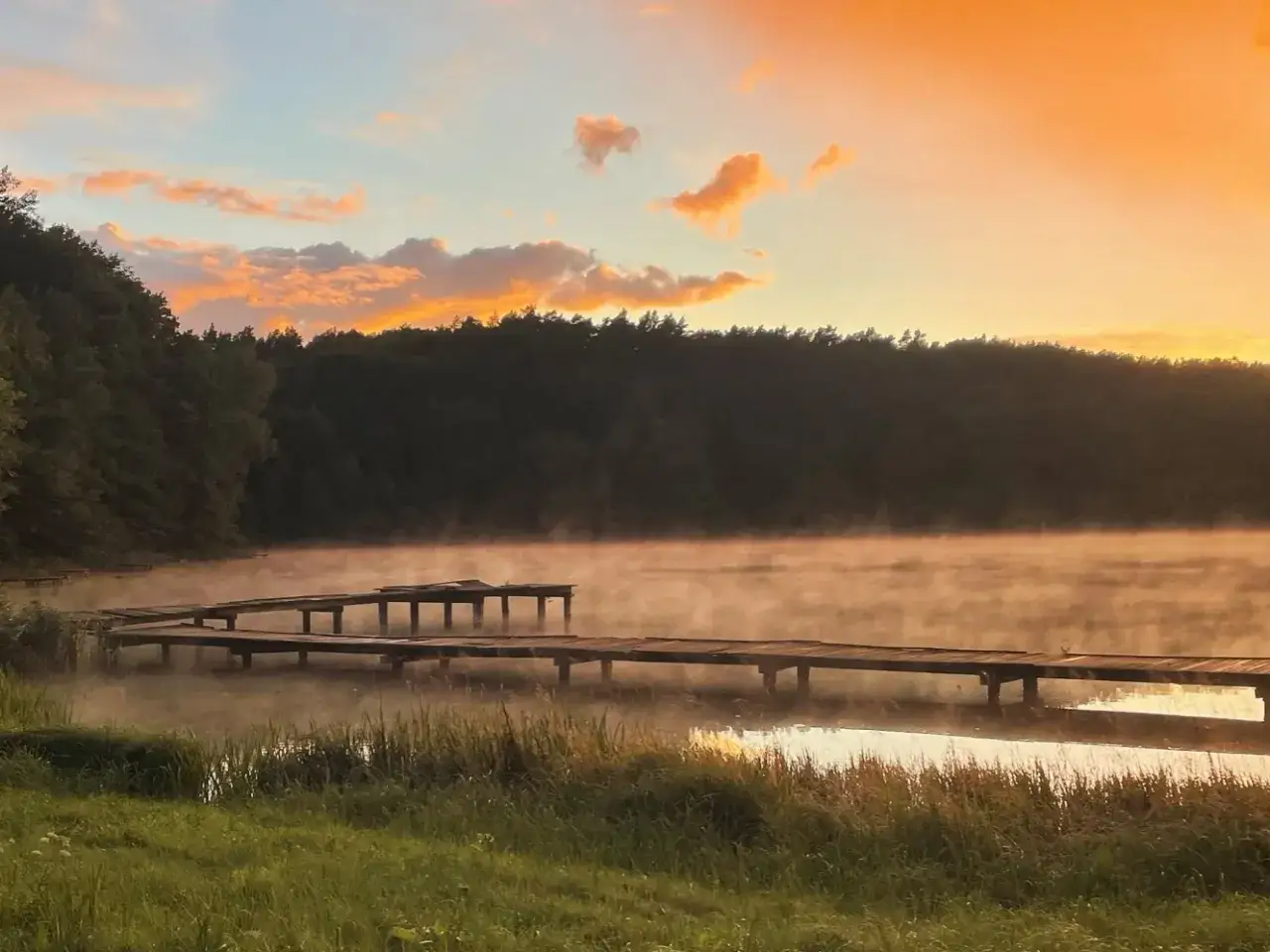Mglisty poranek nad jeziorem w Torzymiu. Drewniane pomosty zanurzają się we mgle, a niebo rozświetla ciepły, pomarańczowy blask.