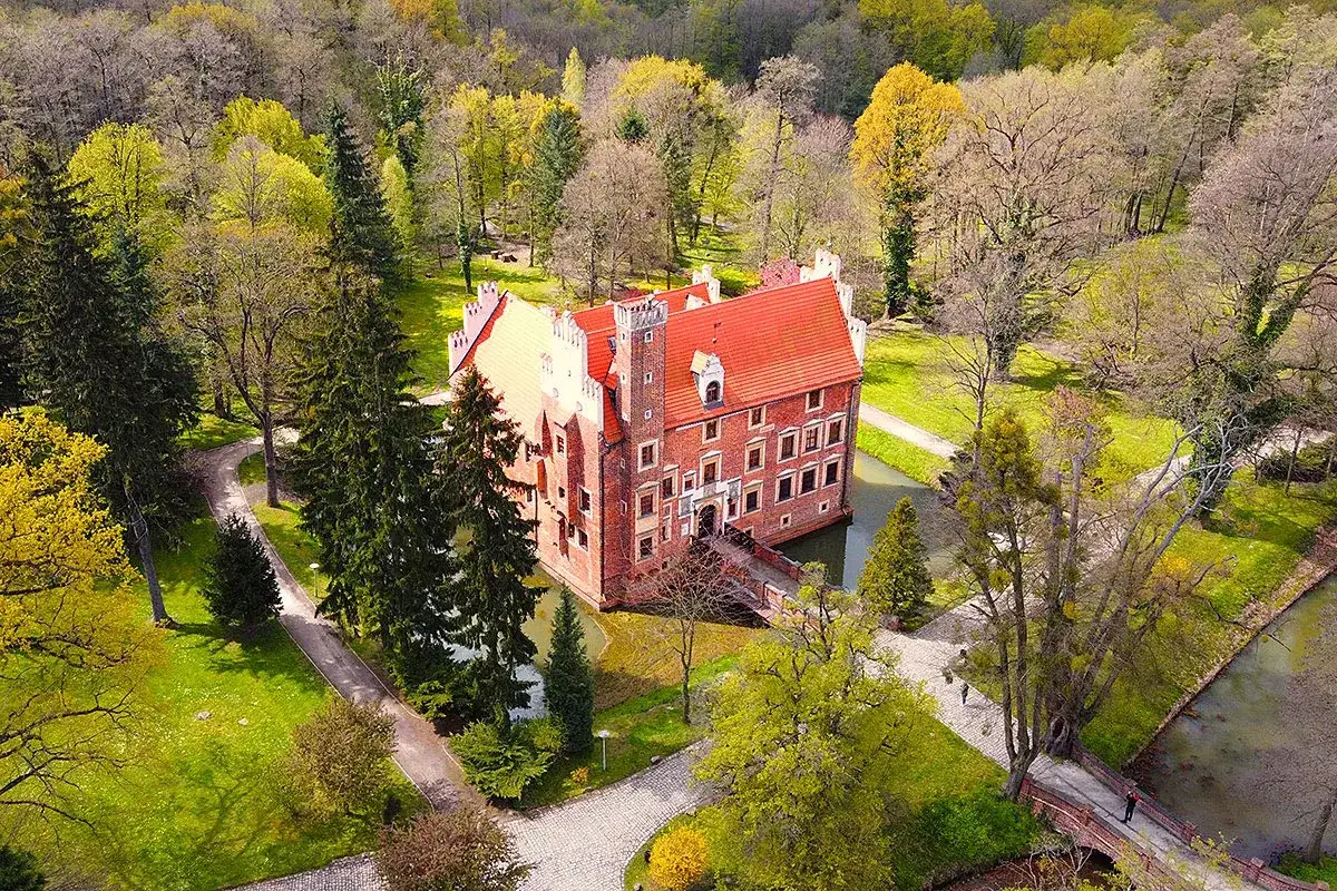 Zamek na wodzie w Wojnowicach otoczony zielenią i drzewami, z czerwoną dachem i ceglanymi murami.