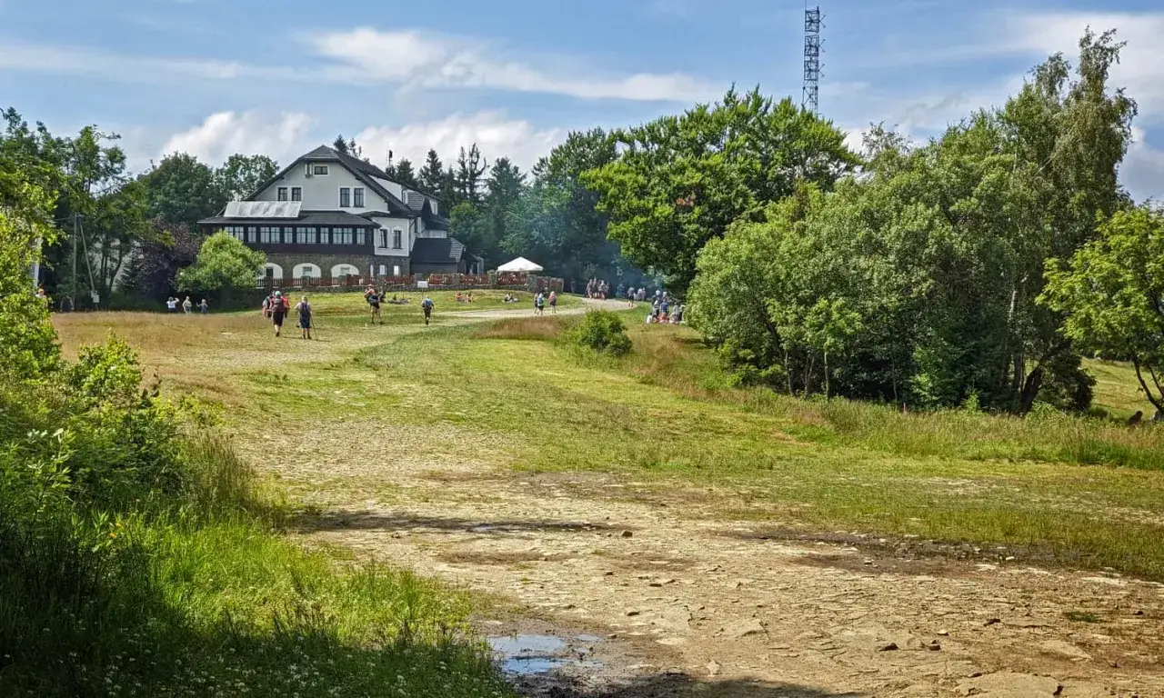 Ludzie spacerują wokół schroniska na Magurce Wilkowickiej. W tle las i wieża telekomunikacyjna.