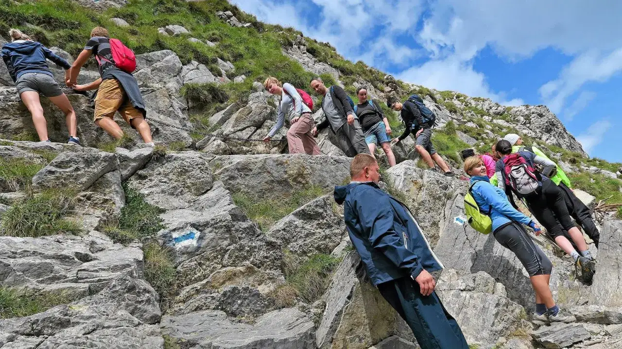 Grupa turystów wspina się po kamienistym szlaku. Widać łańcuchy i niebieskie znaki. Czas wejścia na Giewont zależy od kondycji.