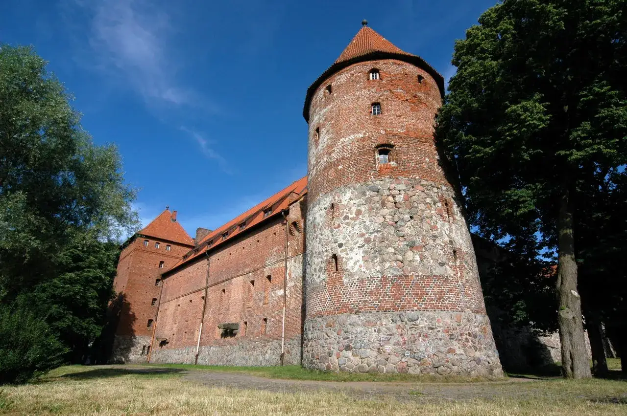 Zamek w Bytowie, potężna ceglana budowla z kamienną podstawą, góruje na tle błękitnego nieba. Otoczony zielenią, zaprasza do odkrycia swojej historii.