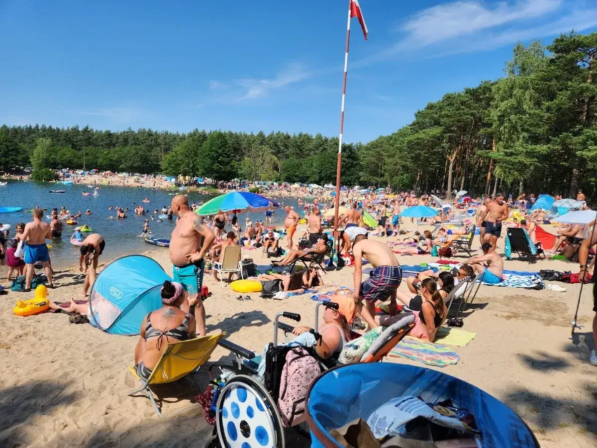 Tłum ludzi odpoczywa na plaży nad jeziorem, wśród nich osoby na wózkach. W tle las, a na wodzie mnóstwo pływających. Wszędzie widać kolorowe parawany i ręczniki, a także charakterystyczne dla lata rokitki.